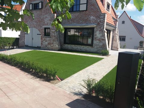 Voortuin met kunstgras, kasseipad en jonge taxushaag bij een villa in Duinbergen