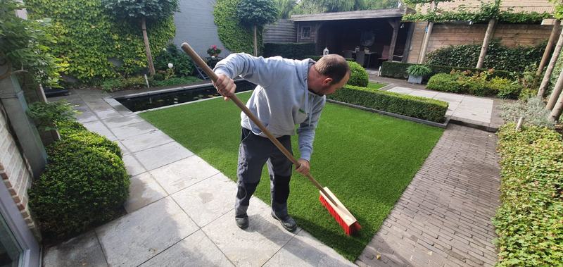 Medewerker van ForGrass borstelt pas gelegd kunstgras op met een harde bezem, naast een vijver in Eeklo